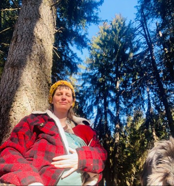 Oli sitz im Wald in der Sonne. Oli hat braune Haare und trägt eine gelbe Kappe und eine rot schwarz karrierte Jacke. Oli hält ein Kleinkind mit einer roten Mütze auf. Im rechten unteren Rand sieht man die Schnautze von einem grauen Hund.