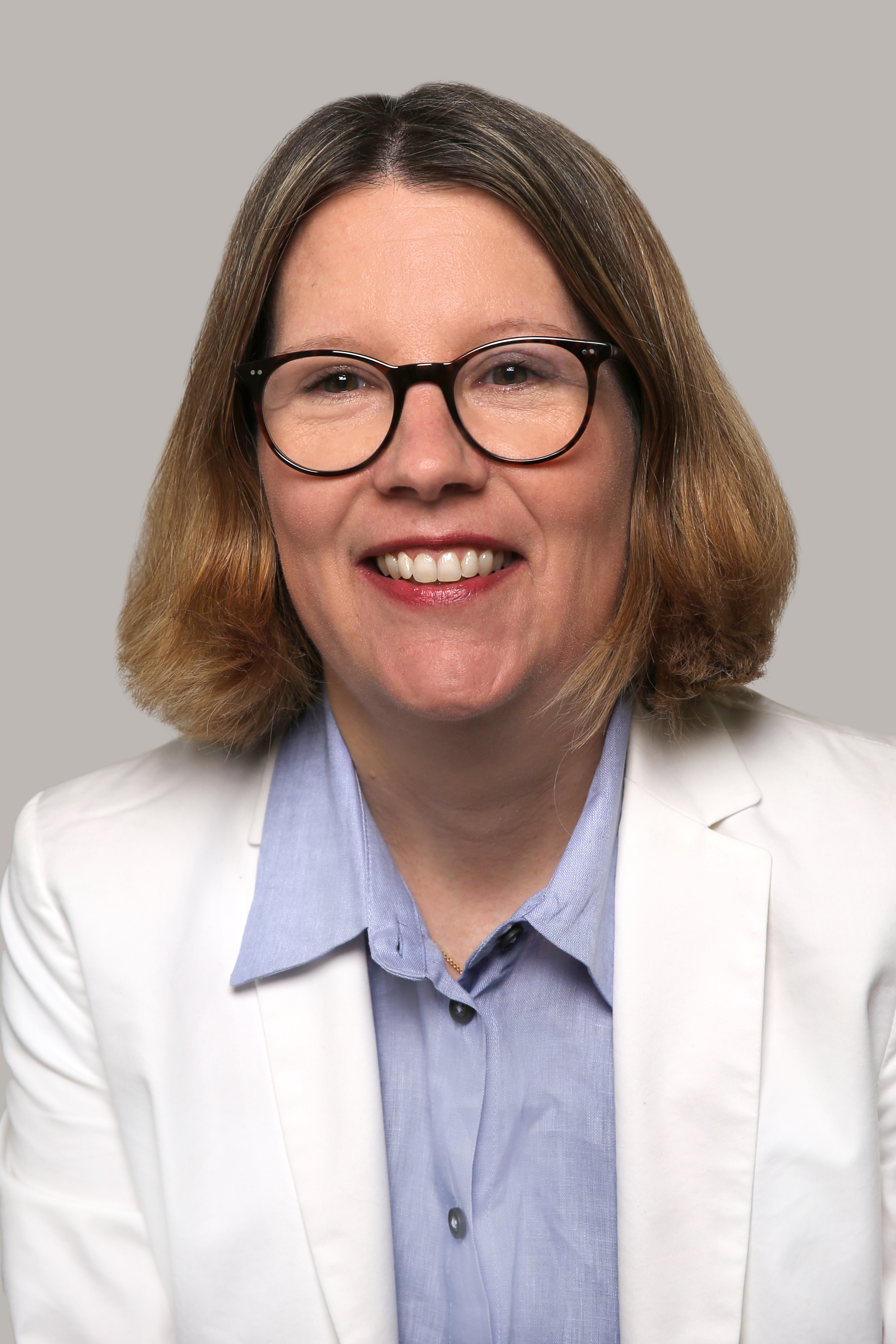 Karin Huber hat kurze dunkelblonde Haare, trägt eine schwarze Brille, ein blaues Hemd und ein weisses Jacket und lacht in die Kamera