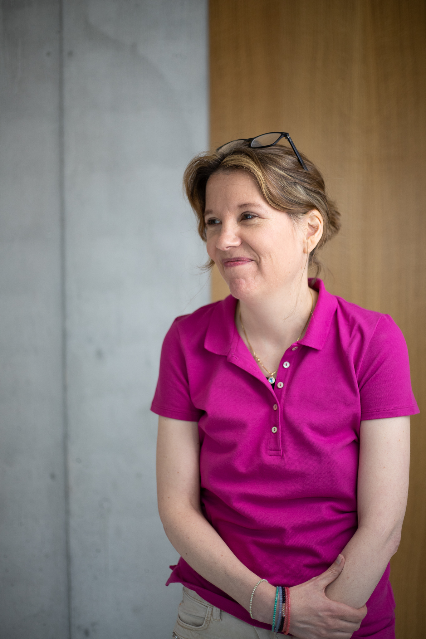 Karin lächelt und blickt abseits der Kamera, sie trägt ein pinkes Hemd und auf ihrem Kopf sitzt eine schwarze Brille