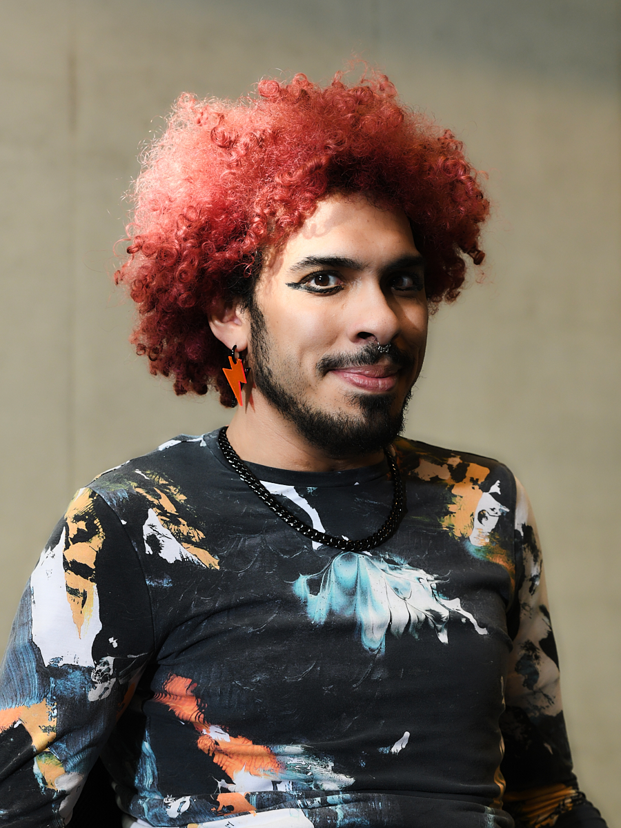 Edwin ist eine schwarze Person mit Bart, mit pinkem Afro, Eyeliner und orangen Blitz-Ohrenringen, schwarzem ,weissem und orangen Shirt.