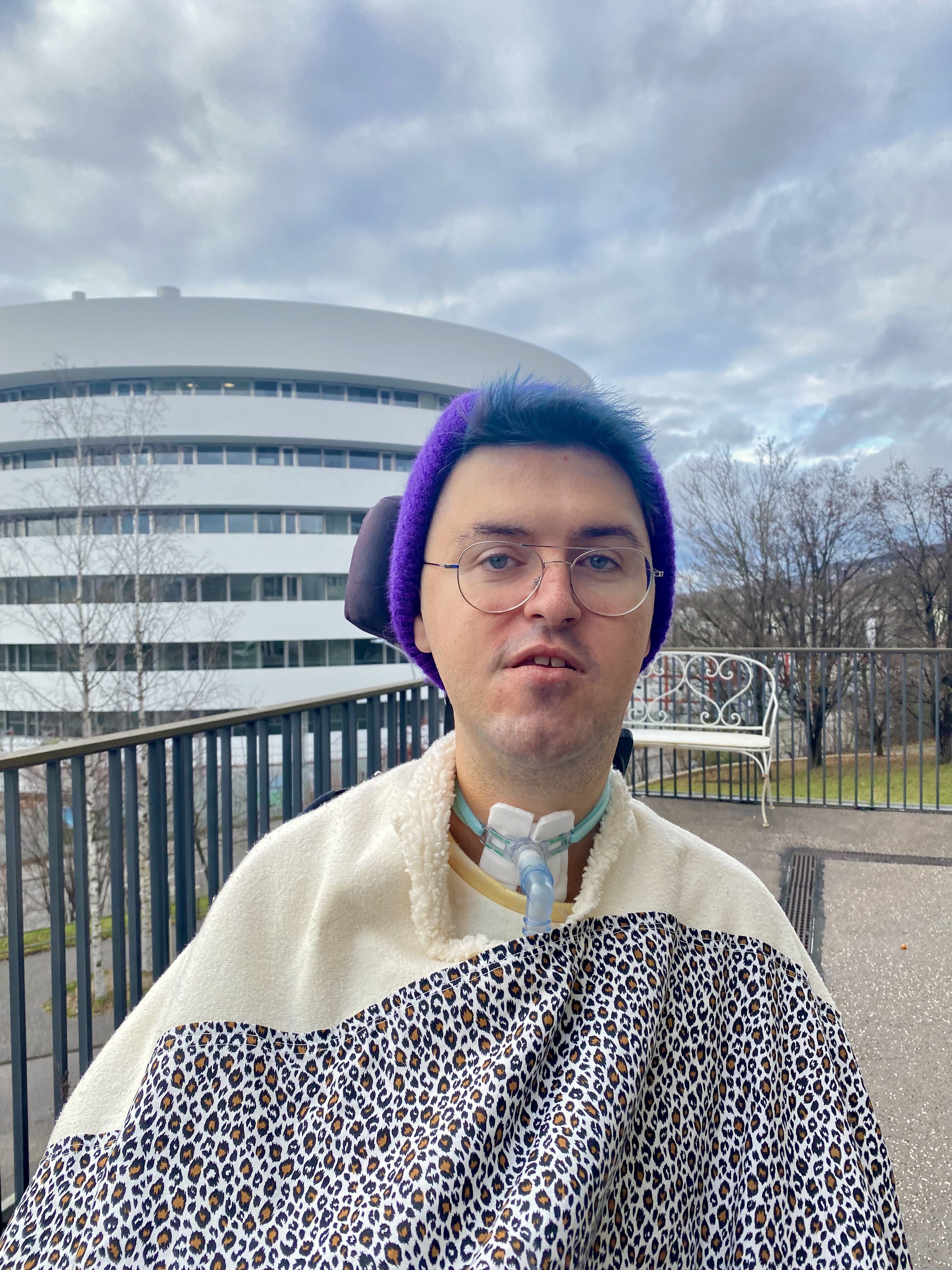Foto von Lila, mit einer lila Mütze, blauen Haaren, einer Brille und einem weissen Gewand. Im Hintergrund sind Wolken und ein Wohngebäude zu sehen.